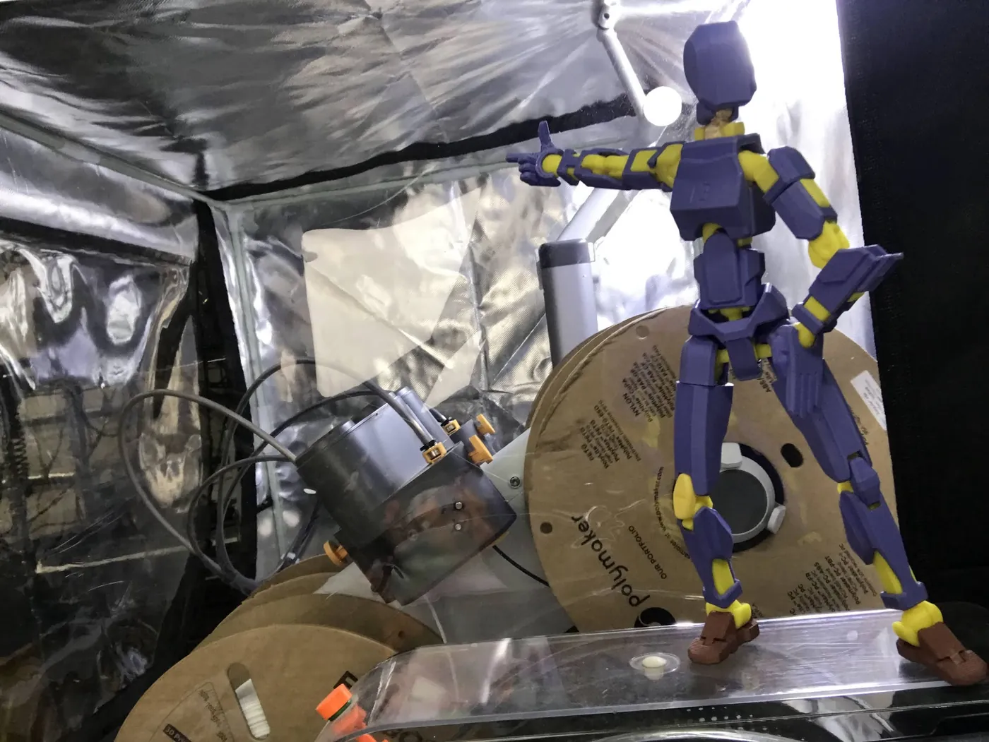 A purple and yellow 3D-printed Dummy Doll pointing, posed on a filament spool inside a reflective print enclosure